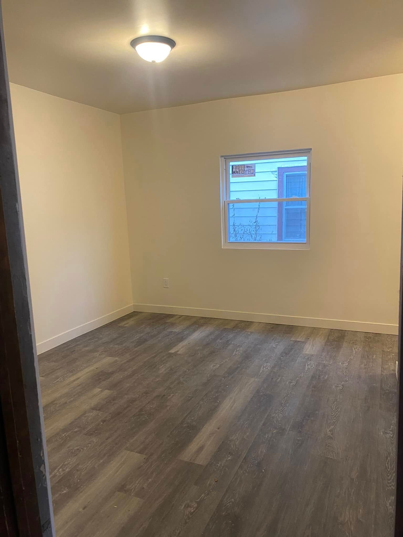 Empty beige room with dark wood flooring, a ceiling light, and a small window.