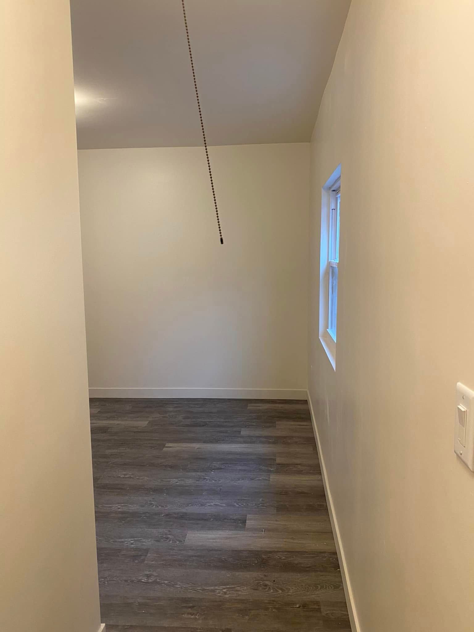 Empty narrow hallway with beige walls, dark wood floor, and a window on the right.
