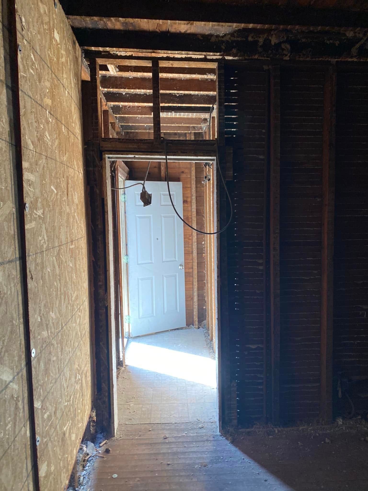 Dim interior hallway with exposed framing and a white door opening into bright sunlight