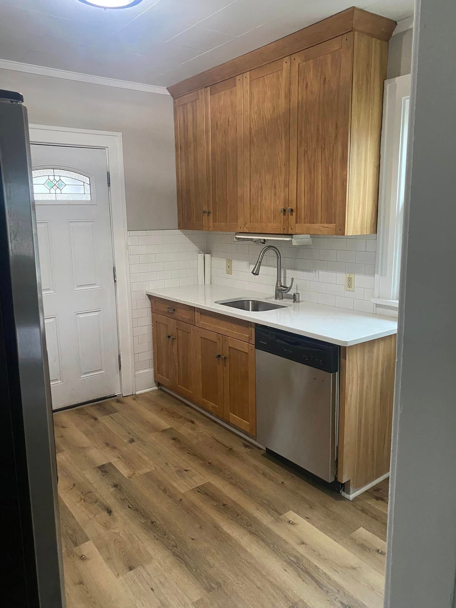 Small kitchen with wood cabinets, white tile backsplash, stainless dishwasher, and a white door.
