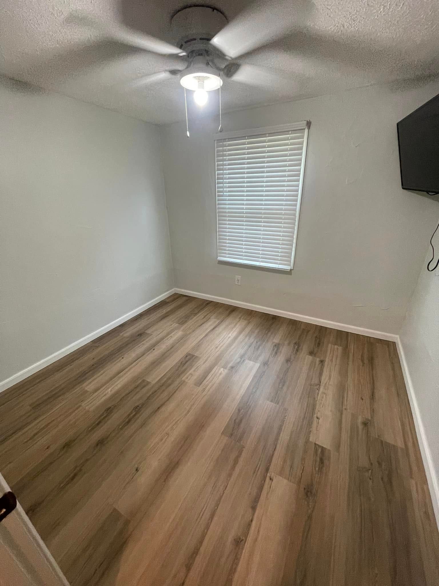 Empty bedroom with wood floors, white walls, ceiling fan, and a window with blinds.