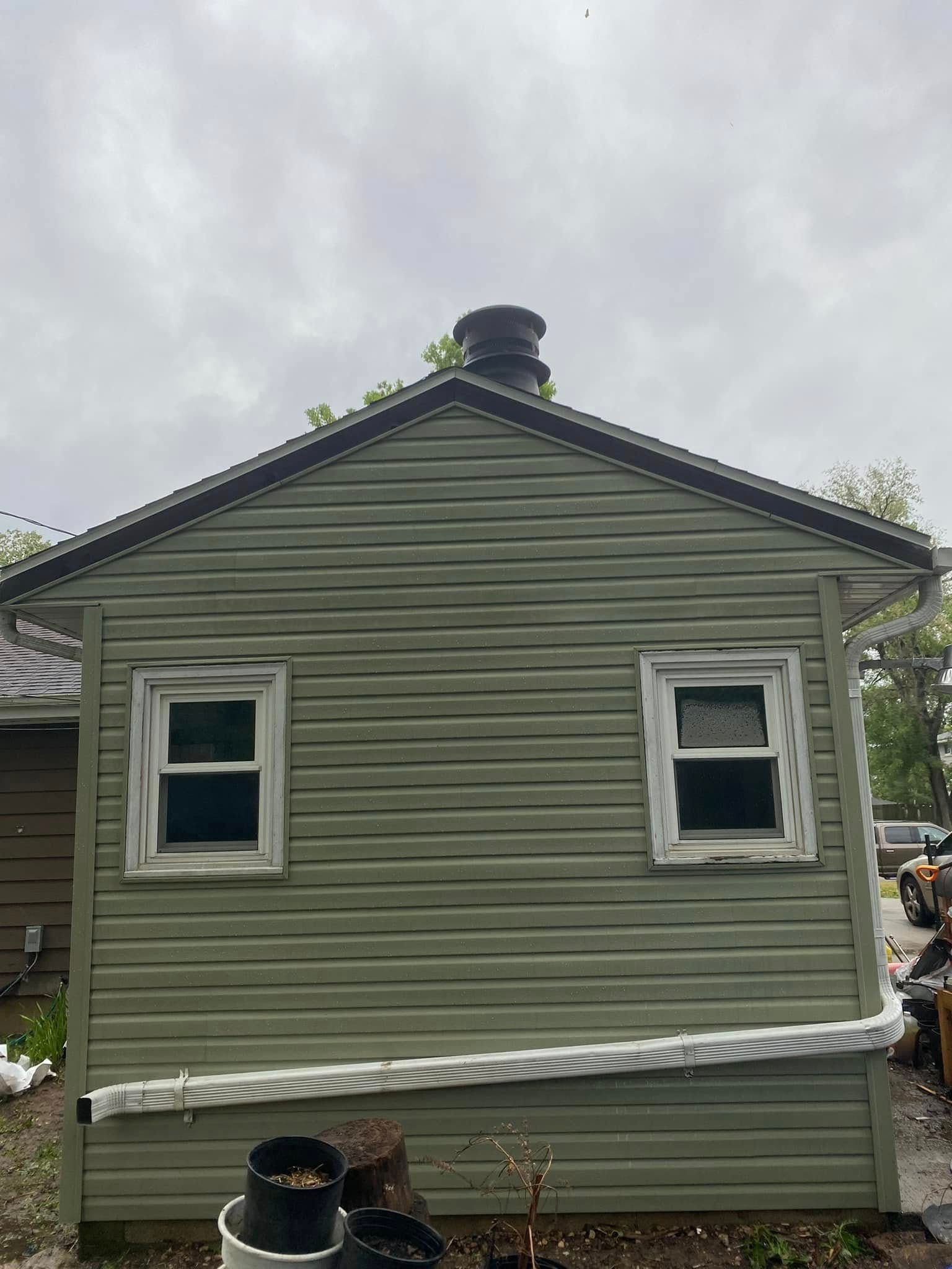Green house side with two windows, small chimney, and tarps along the base under an overcast sky