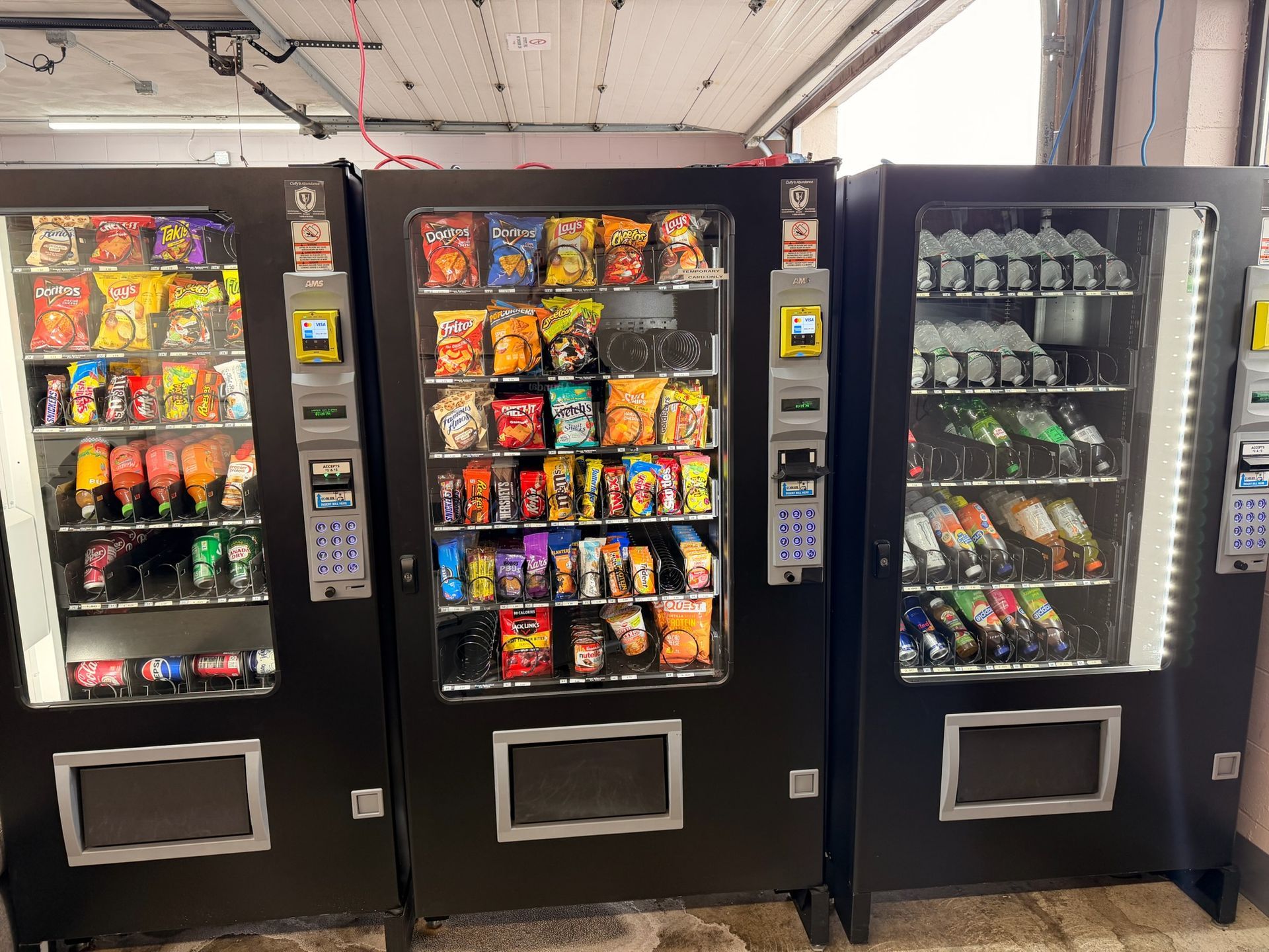 Two white vending machines filled with drinks, standing outside a storefront beside a green dispenser.