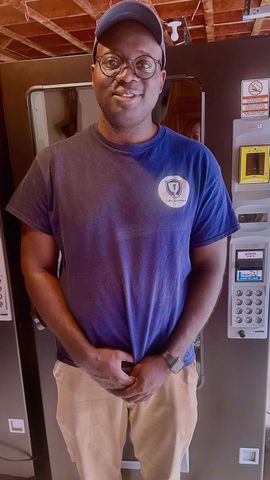 Man standing indoors beside a vending machine, wearing a blue shirt, tan pants, and a cap.