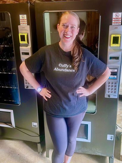 Person standing in front of vending machines, smiling with hands on hips, wearing a dark T-shirt and leggings.