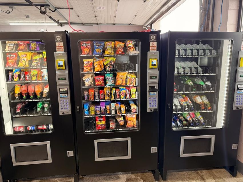 Two white vending machines filled with drinks, standing outside a storefront beside a green dispenser.
