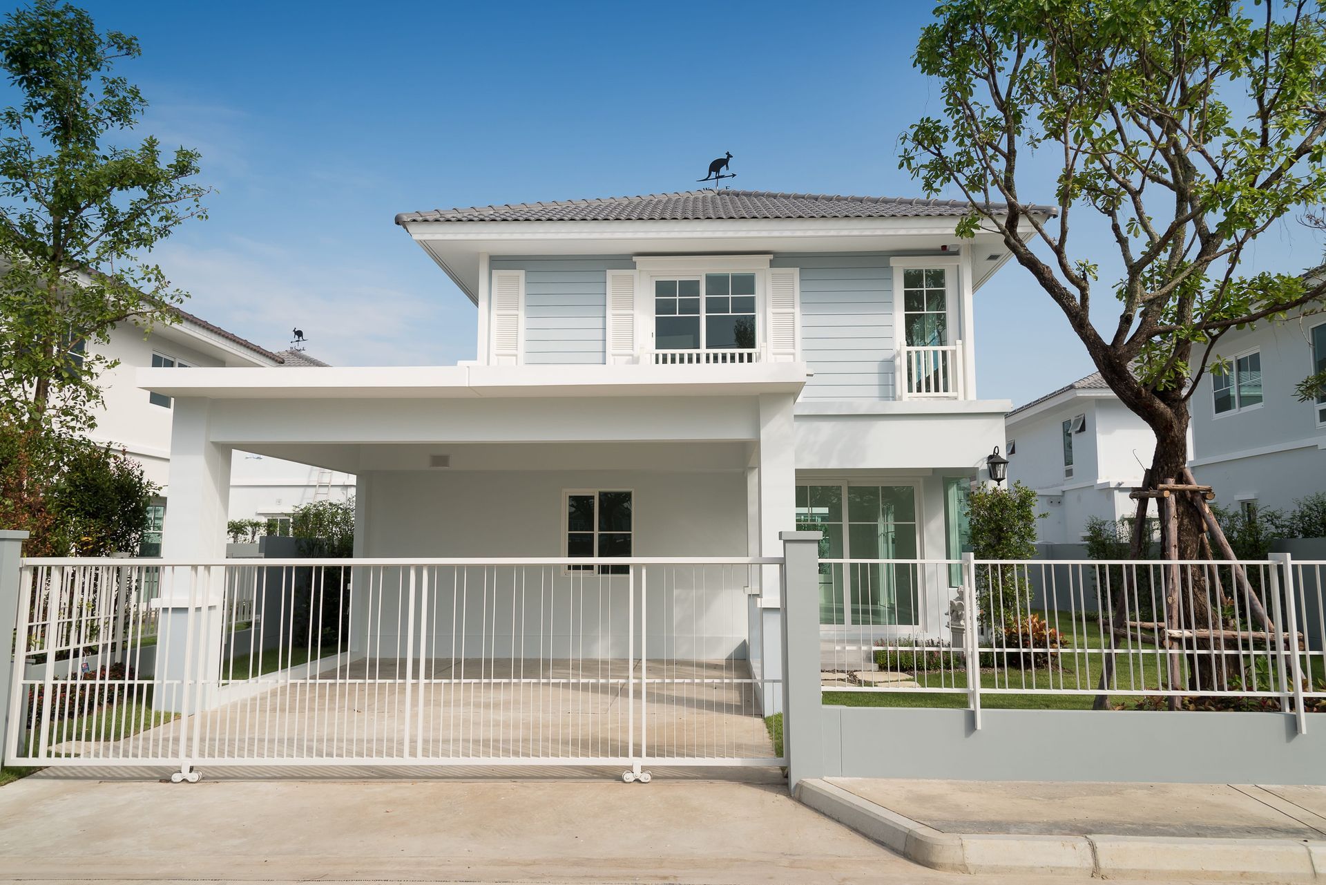 Modern two-story white house with a front gate, driveway, and trees under a clear blue sky