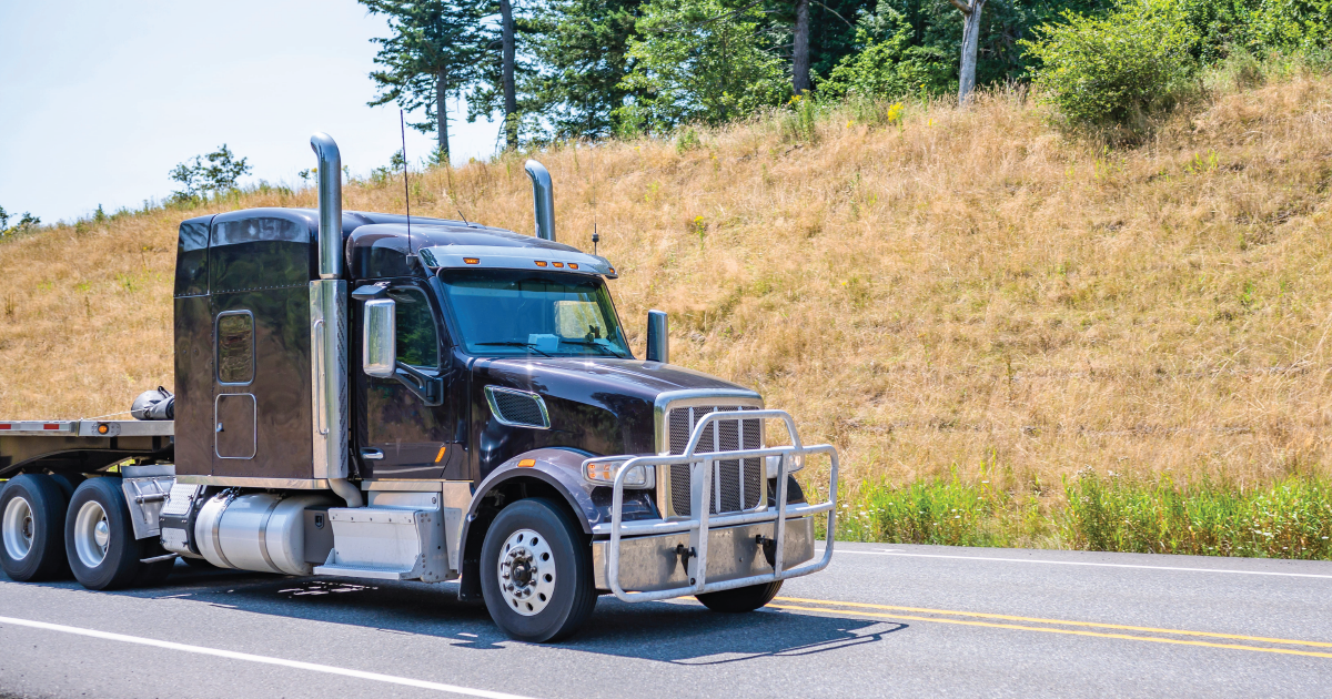 A dark-colored semi-truck with a heavy-duty brush guard driving along a paved road next to a grassy embankment.