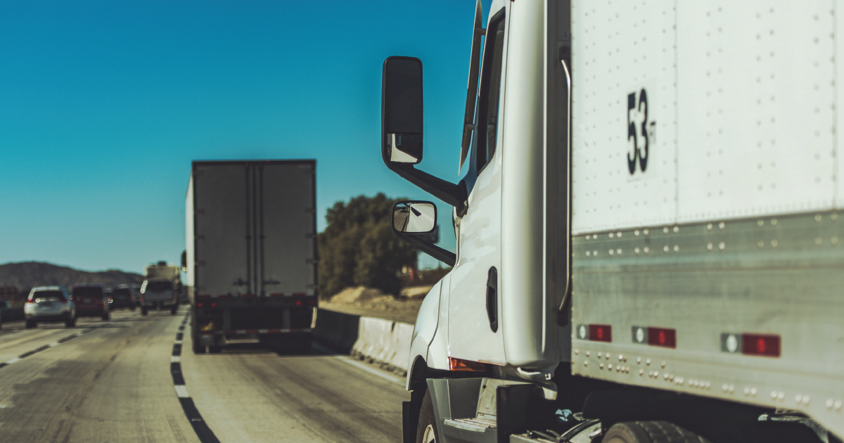 White semi-truck on highway behind a dark truck, blue sky and road barrier visible.
