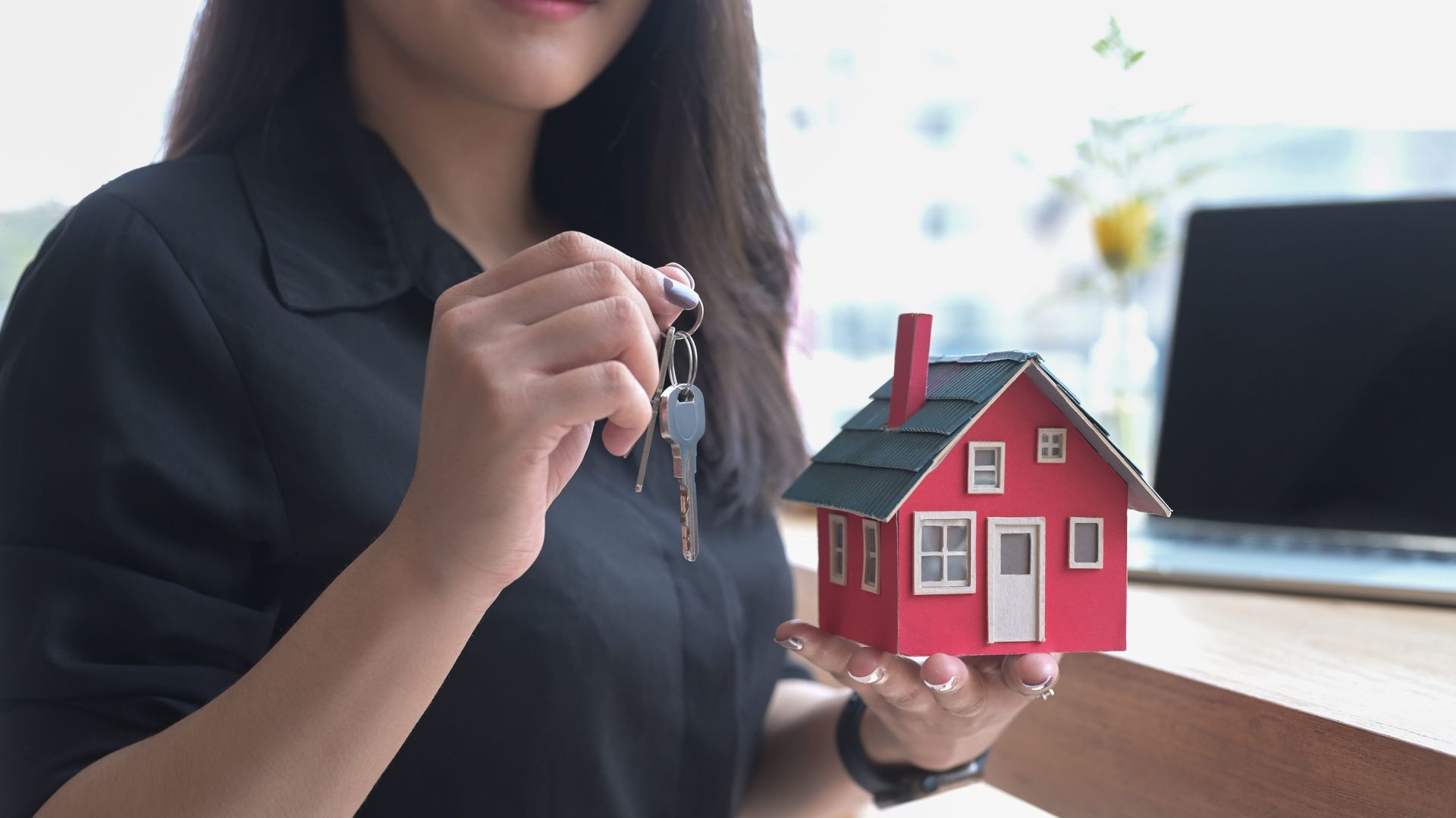 Woman holding house keys beside a red model house on a table, with a laptop in the background