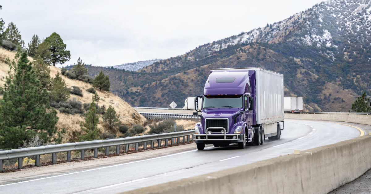 Purple semi-truck driving on a curving mountain highway with snow-dusted hills in the background