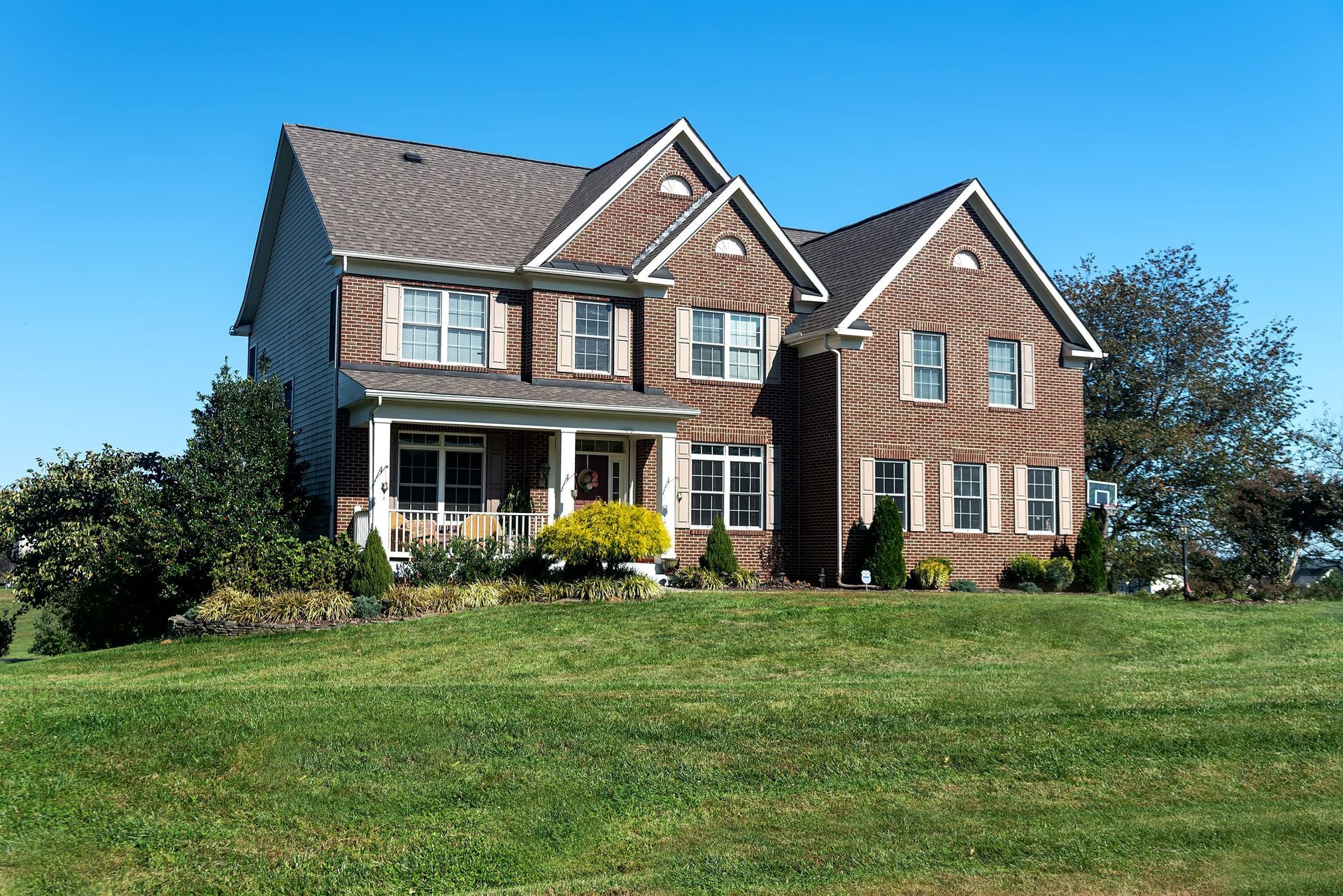 Large brick-and-siding suburban house with a front porch, dormer windows, and a green lawn under a blue sky