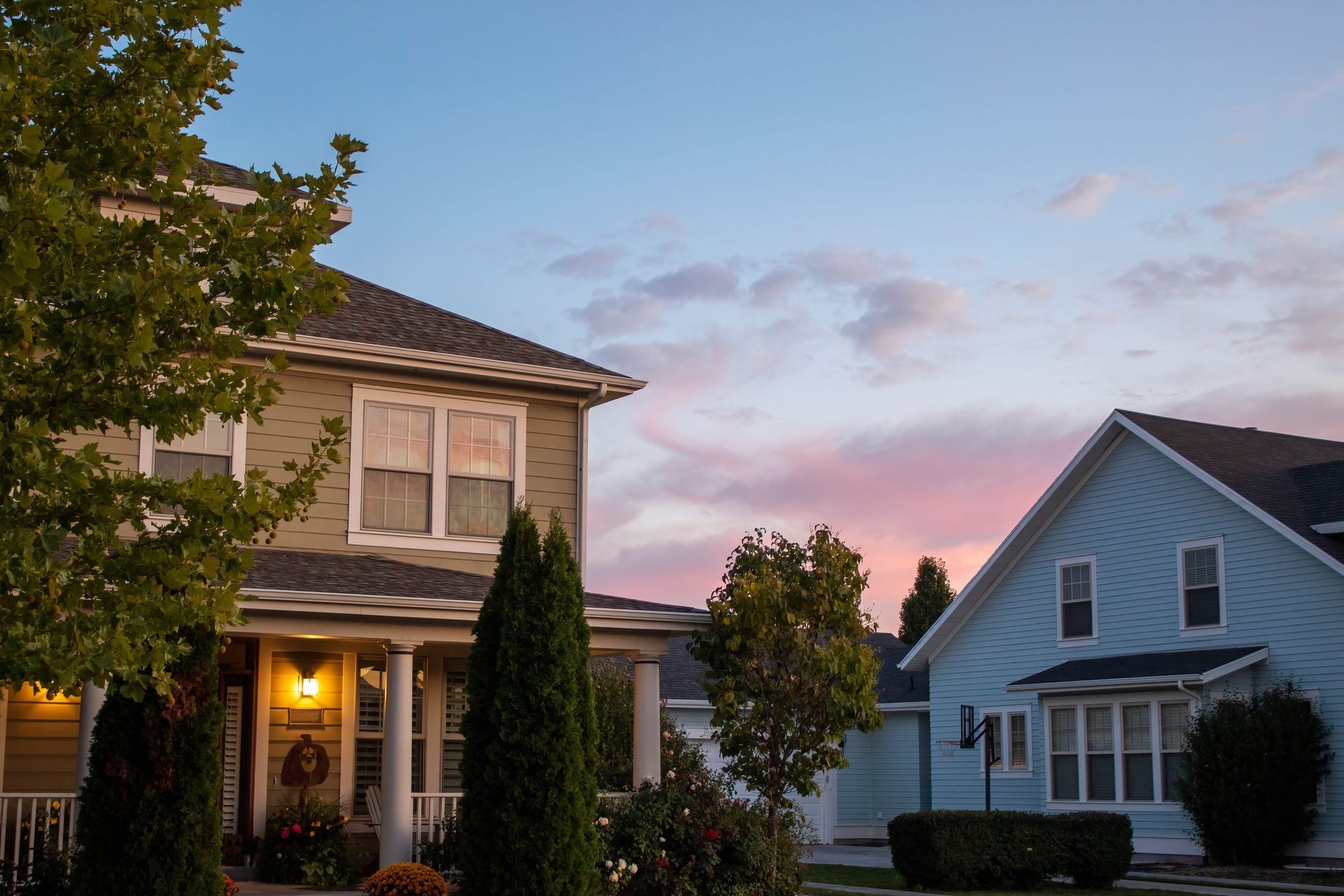 Two suburban houses at dusk, with lit porch on the left and a blue house on the right under a pink sky