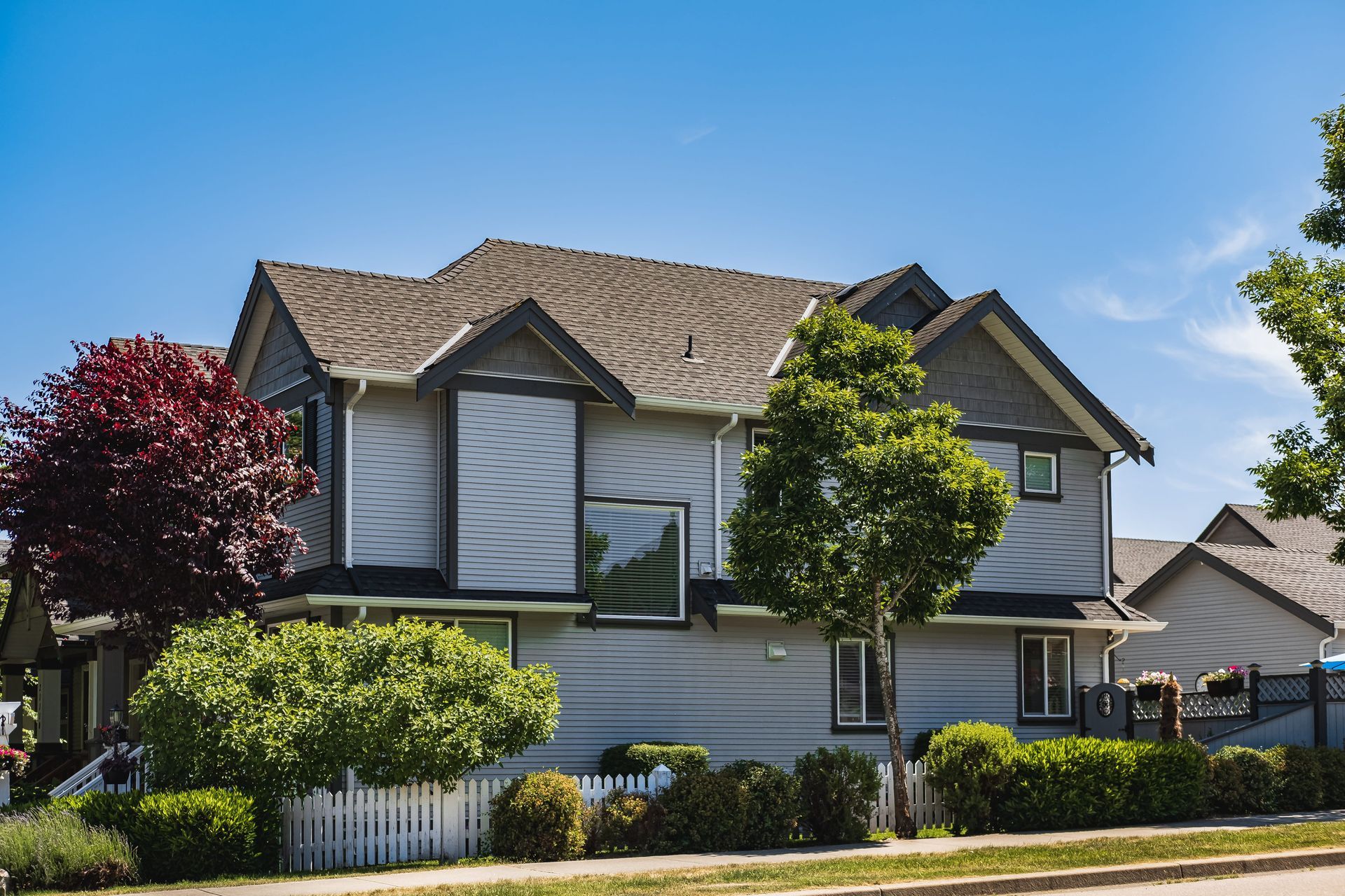 Two-story gray house with white trim, red and green trees, and a white picket fence on a sunny day