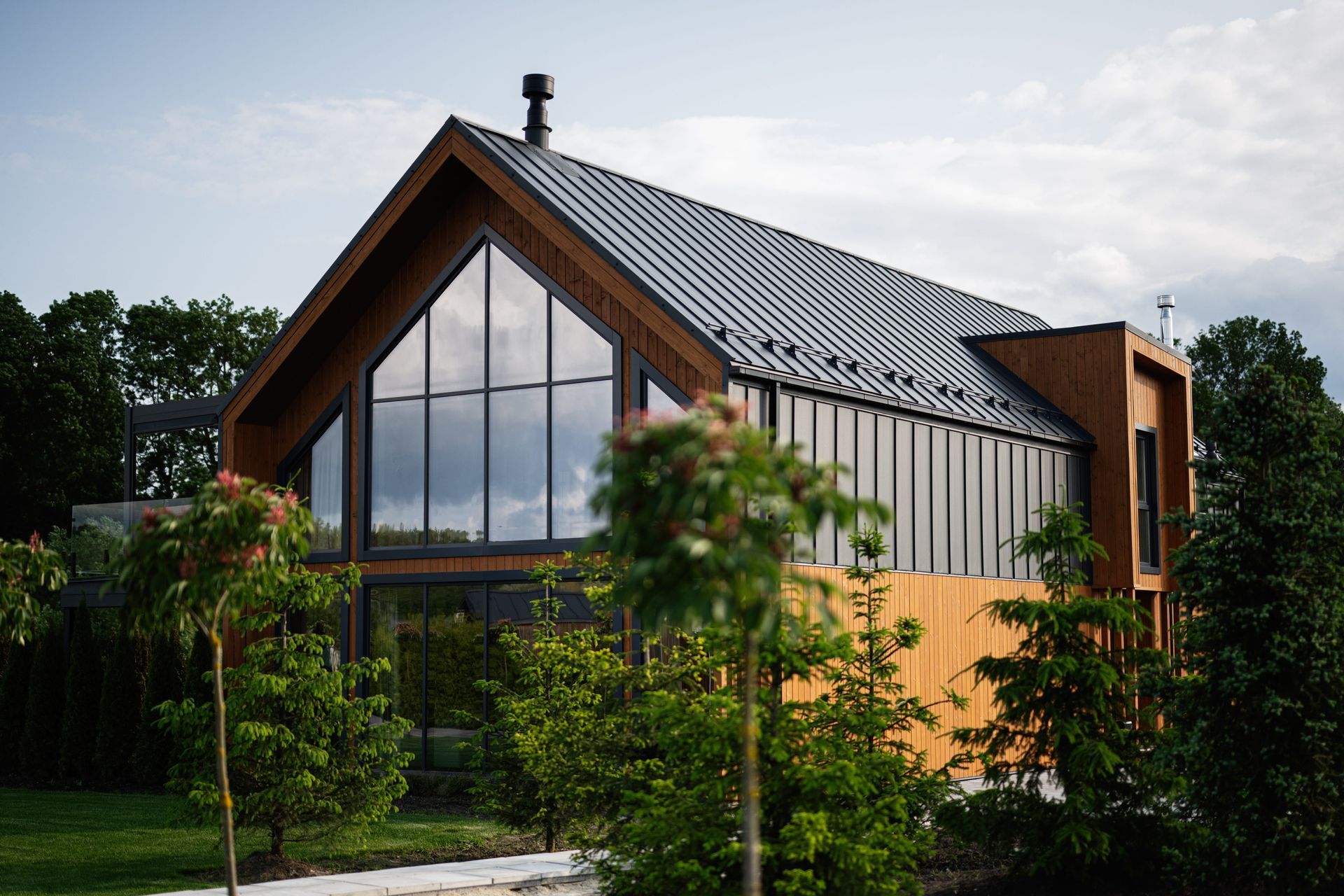 Modern house with large glass windows, dark metal roof, and surrounding green trees