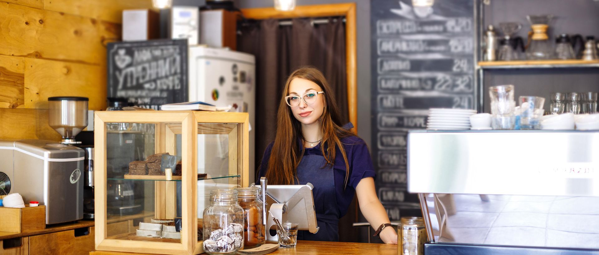 Barista behind a café counter with pastries, shelves, and a white espresso machine