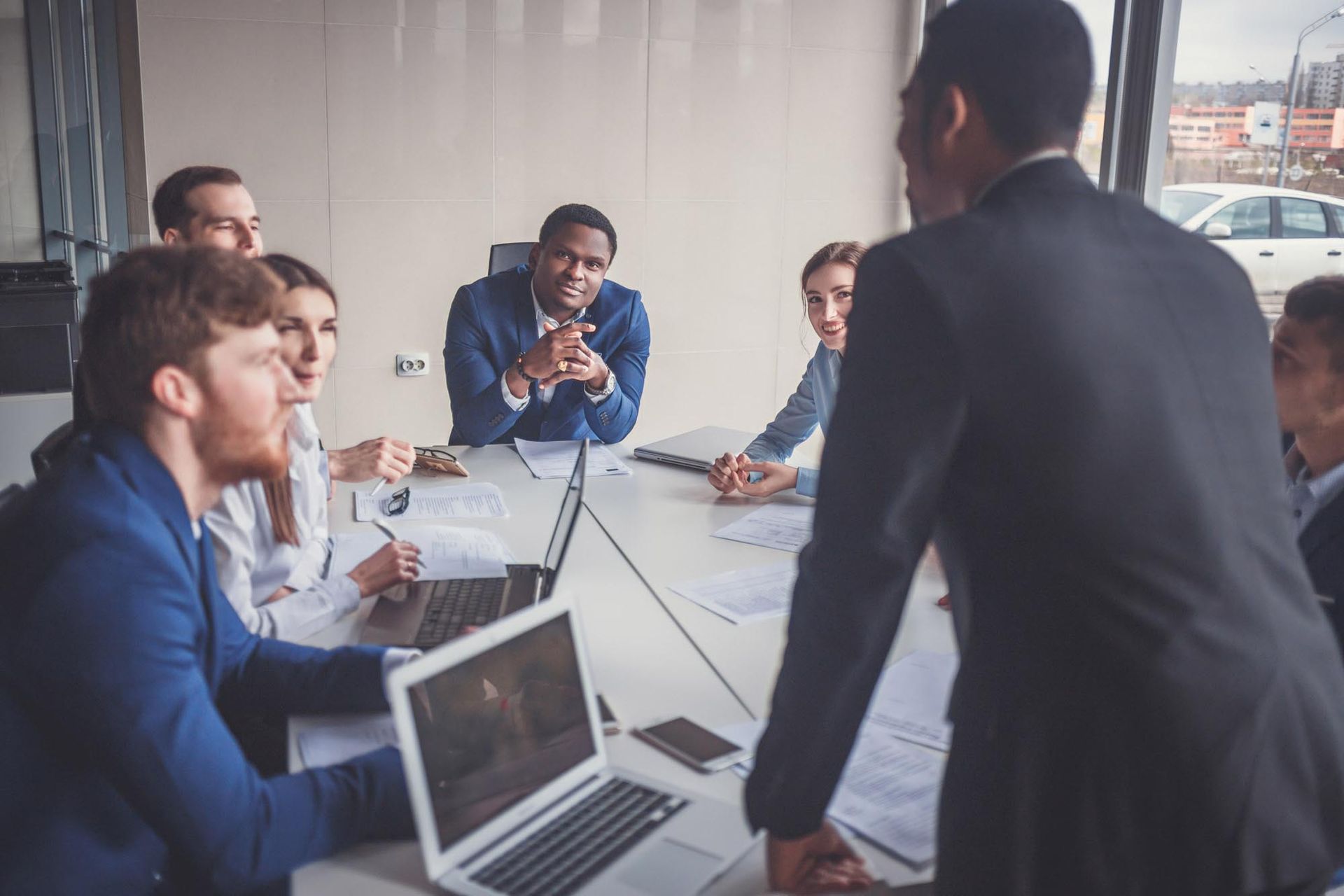 Business meeting around a conference table, with laptops and notes as one person speaks to the group.