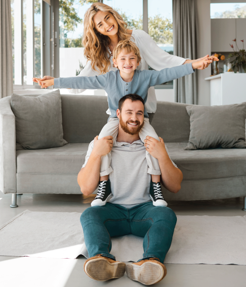 Family playing together in a bright living room, with a child on a man's shoulders and a woman behind them.