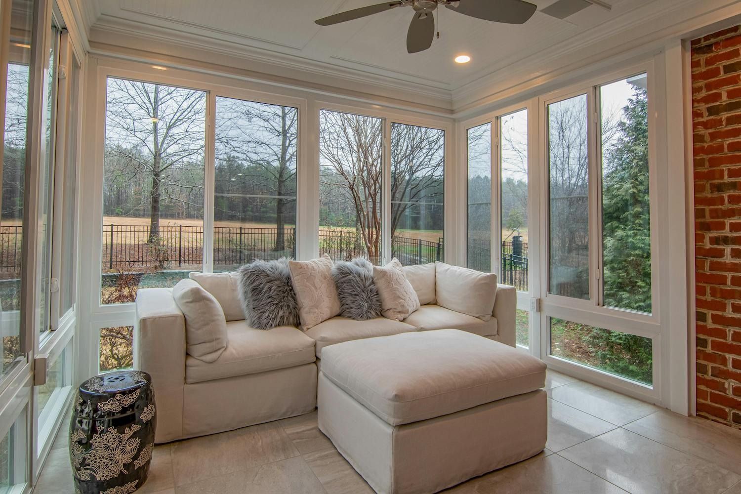 Sunlit sunroom with beige sectional sofa, ottoman, and large windows overlooking trees and yard