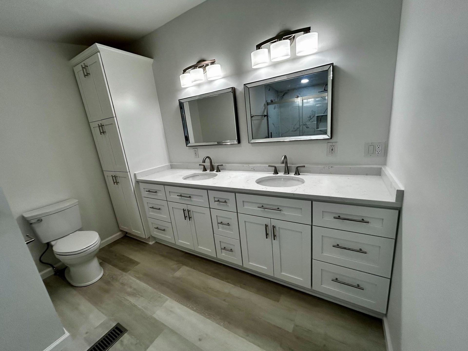 Bright bathroom with double vanity, mirrors, toilet, and white cabinets on wood-look floor.