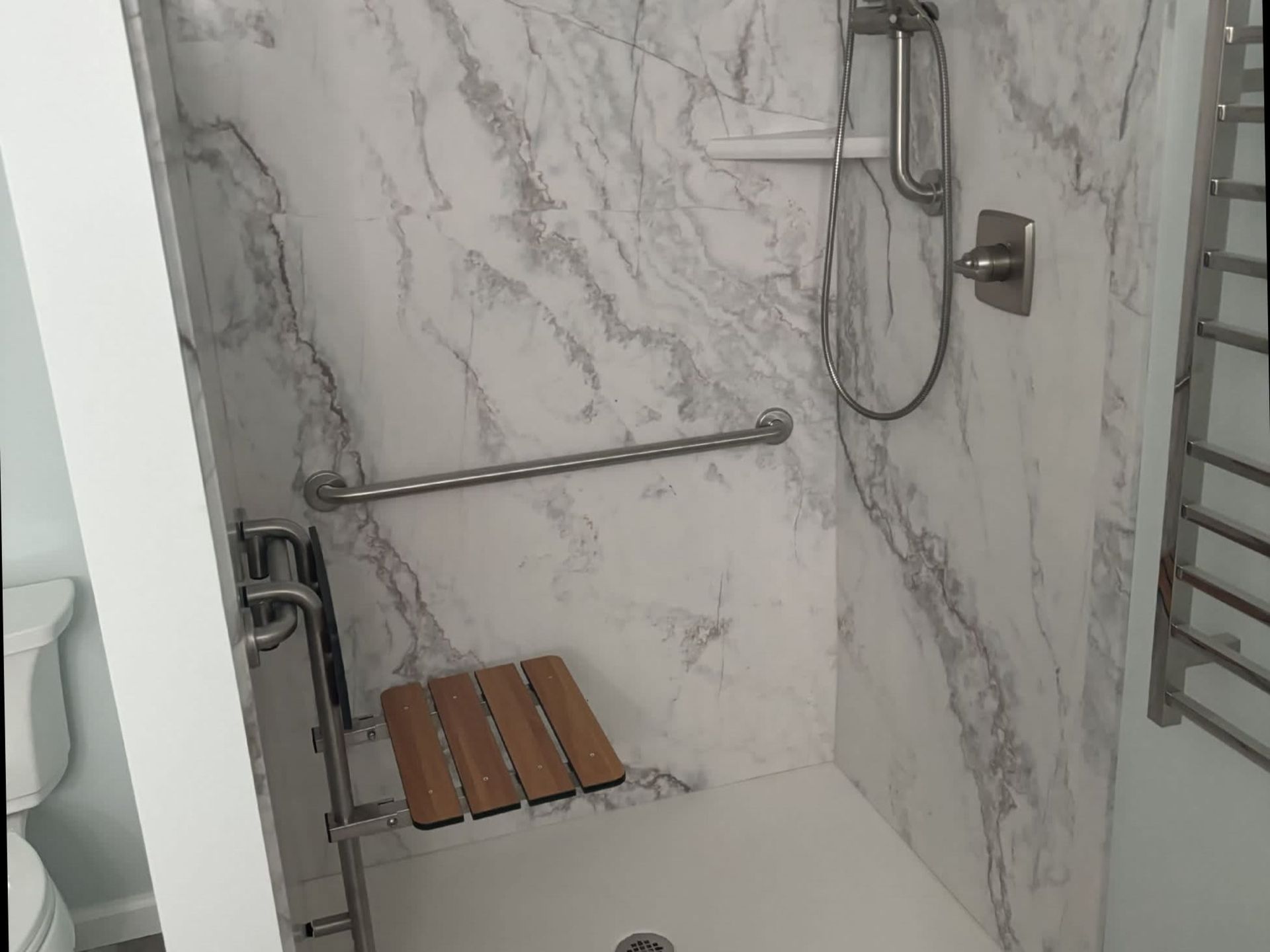 Accessible shower with grab bars, fold-down wooden seat, handheld showerhead, and marble tile walls.