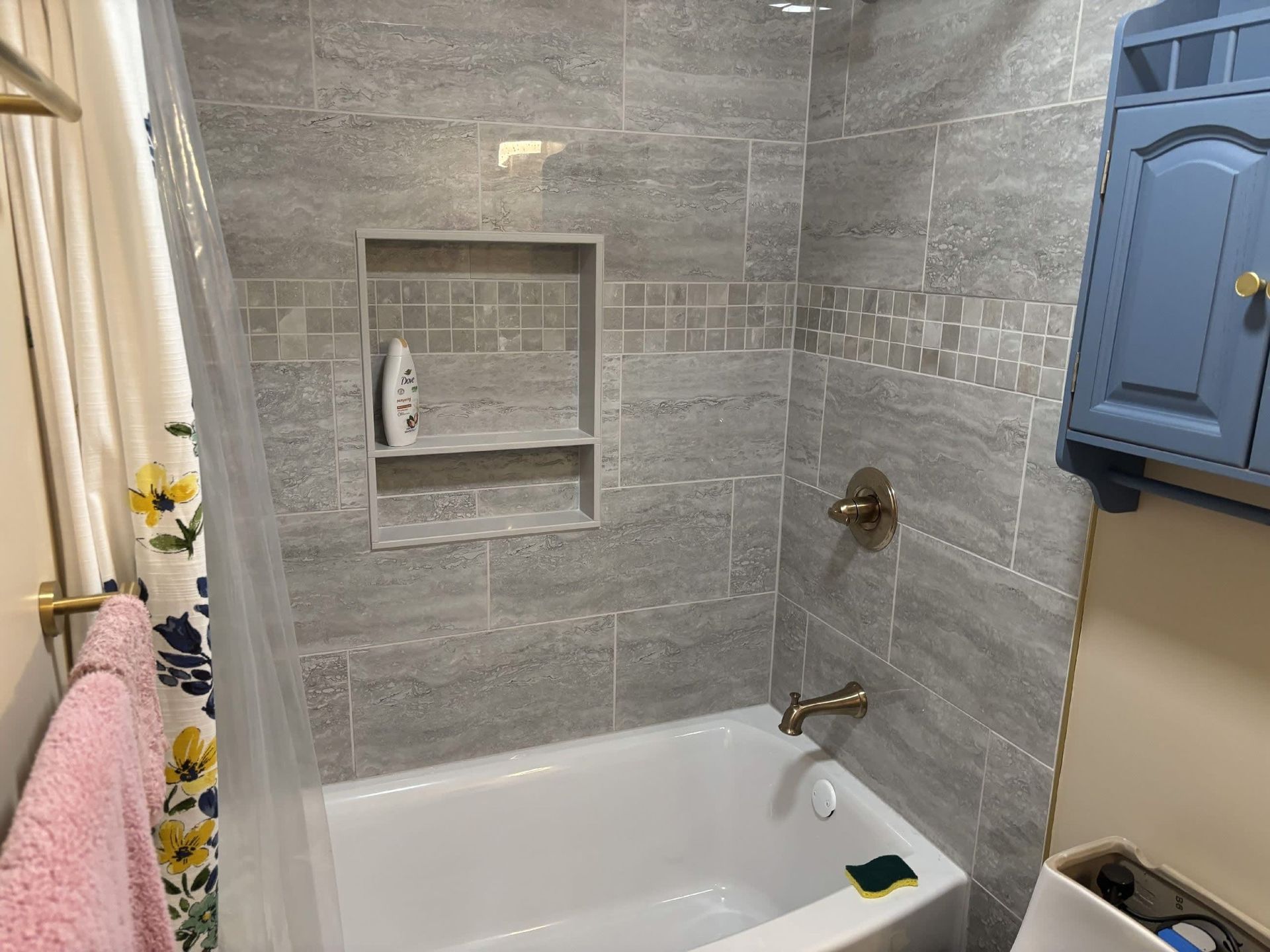 Small tiled bathroom with a white bathtub, built-in shower niche, and blue wall cabinet