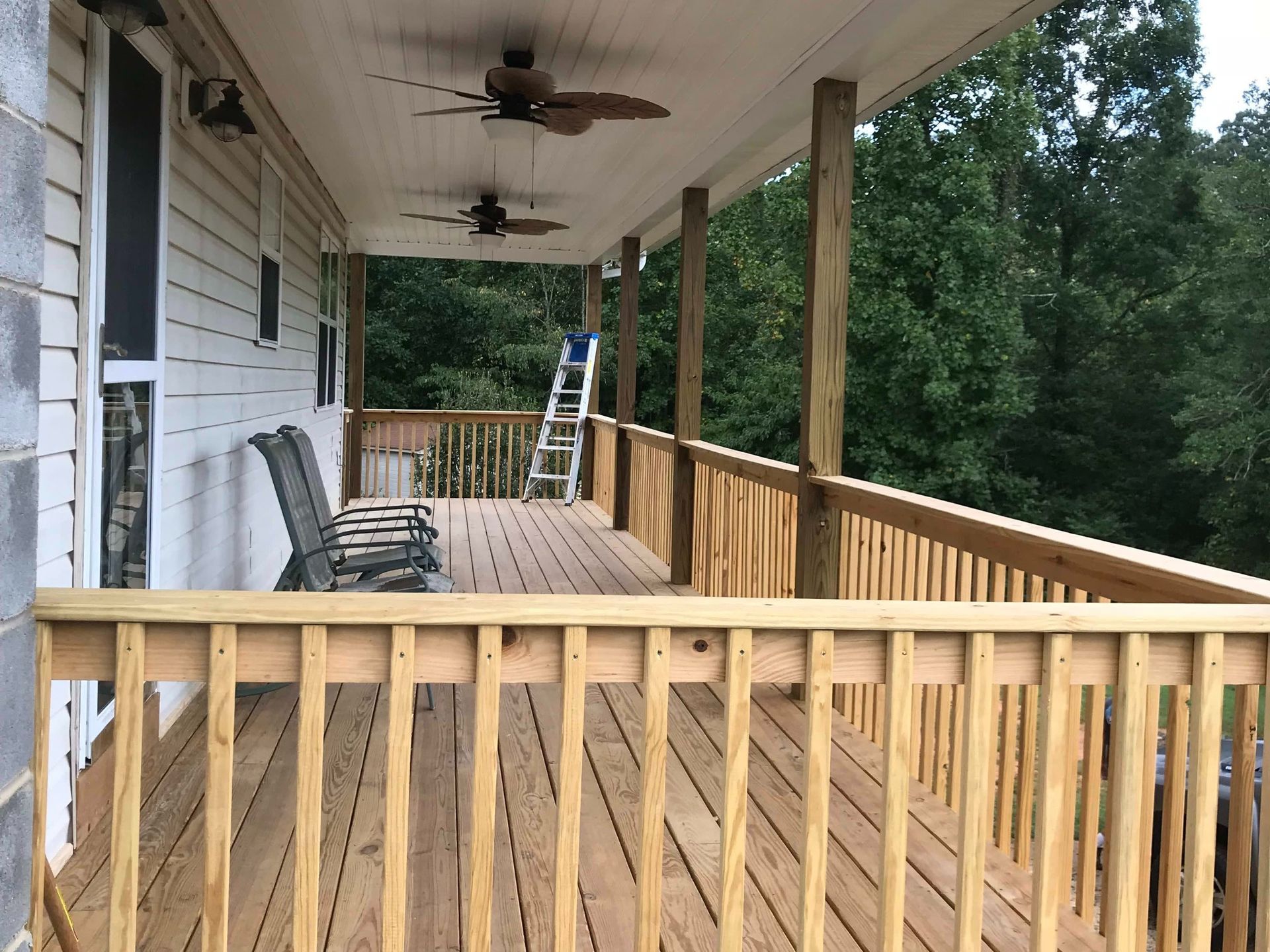Covered wooden porch with railing, ceiling fans, chairs, and a view of trees beyond