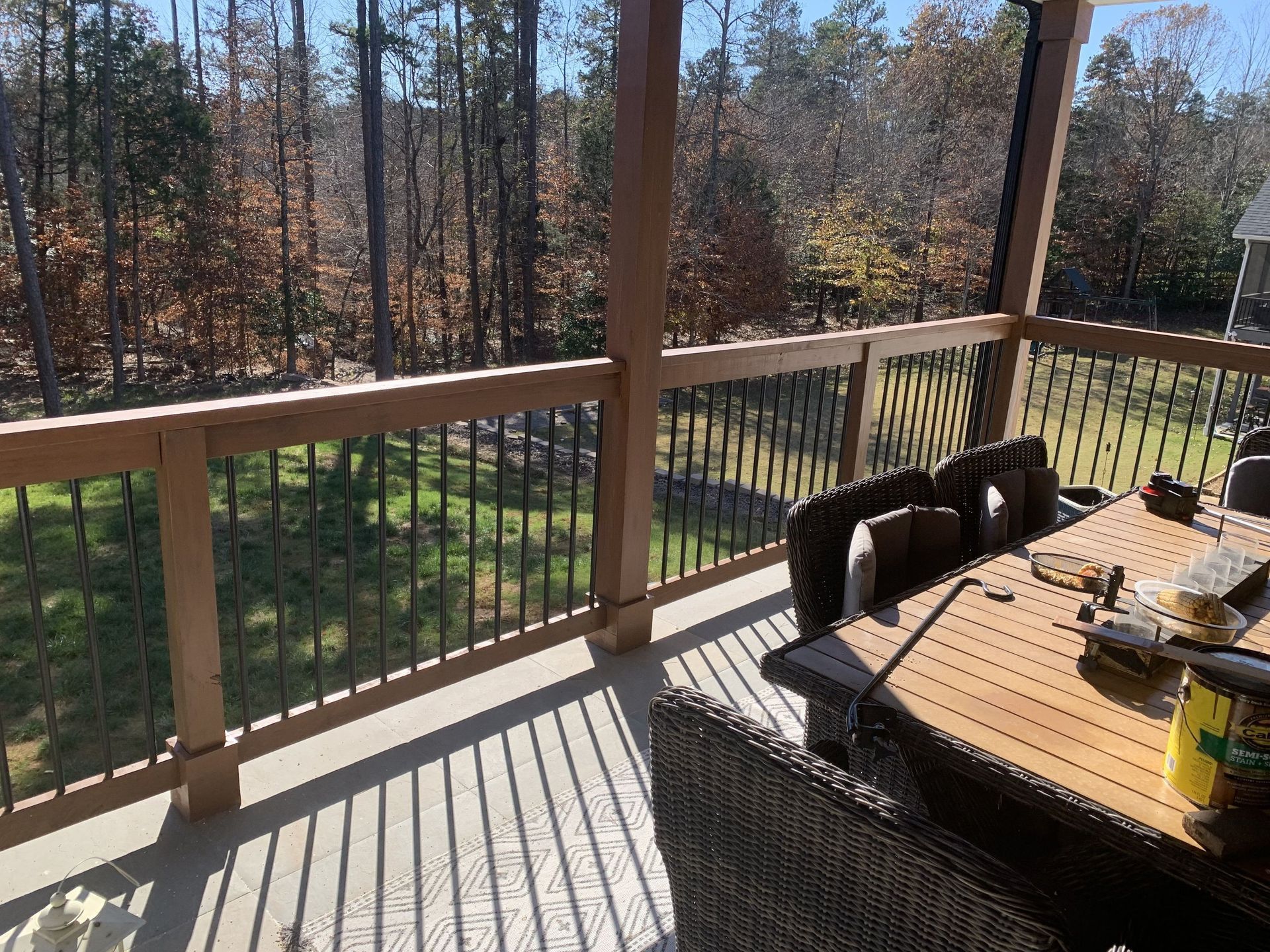 Sunlit backyard deck with wooden railing, trees beyond, and a table with chairs casting long shadows