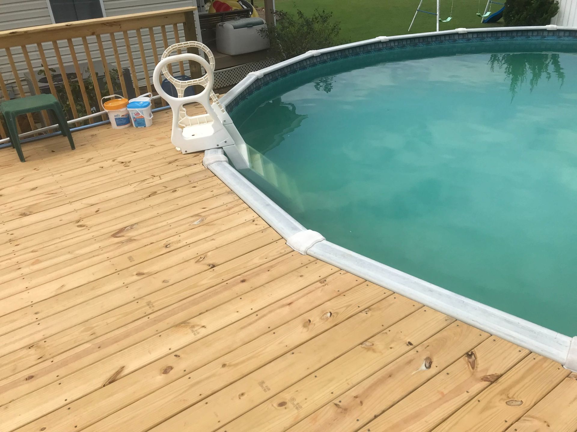 Wooden pool deck beside a turquoise swimming pool with a white railing and chairs.