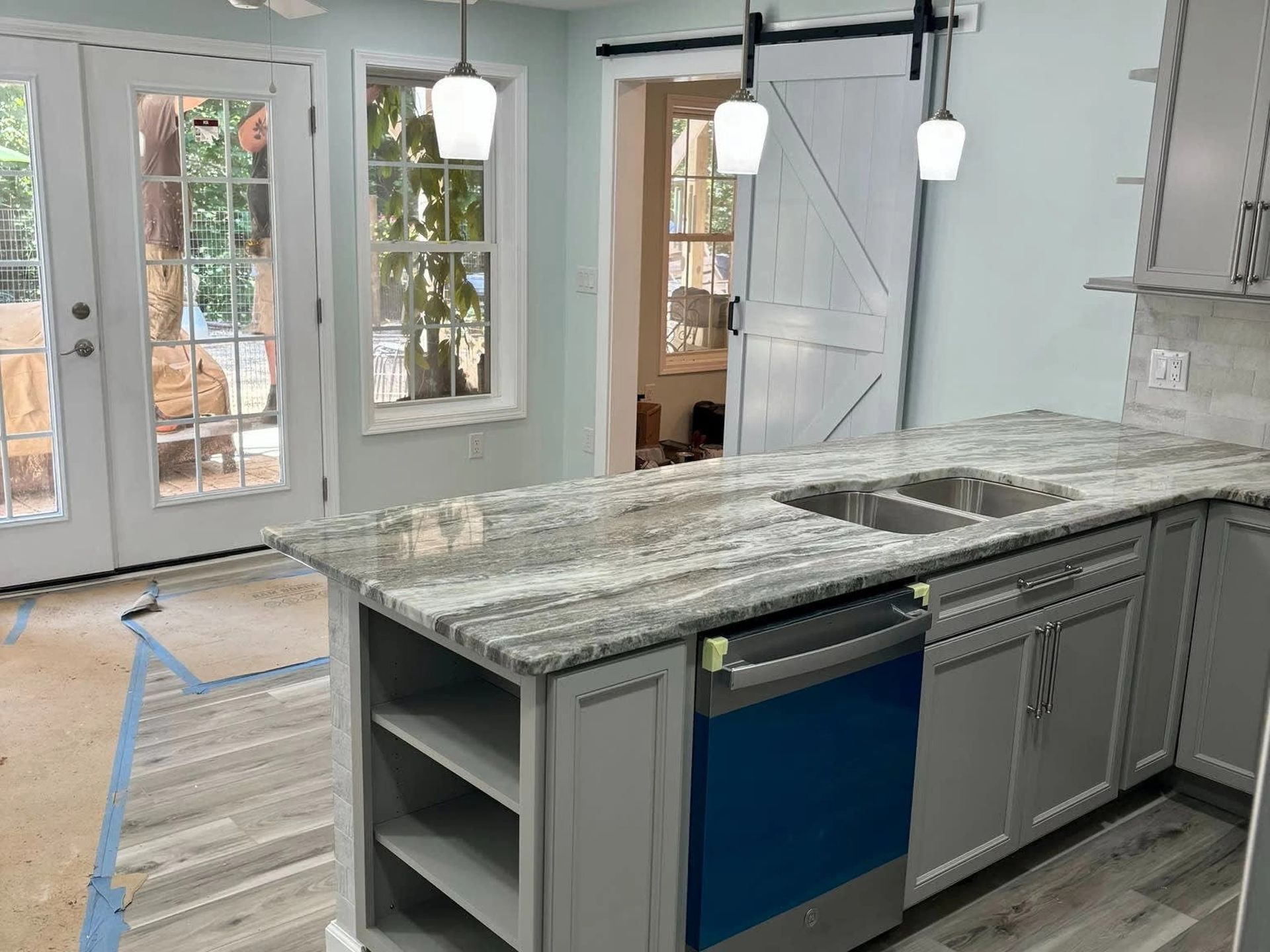 Kitchen under renovation with granite island, gray cabinets, blue dishwasher, and unfinished floorboards