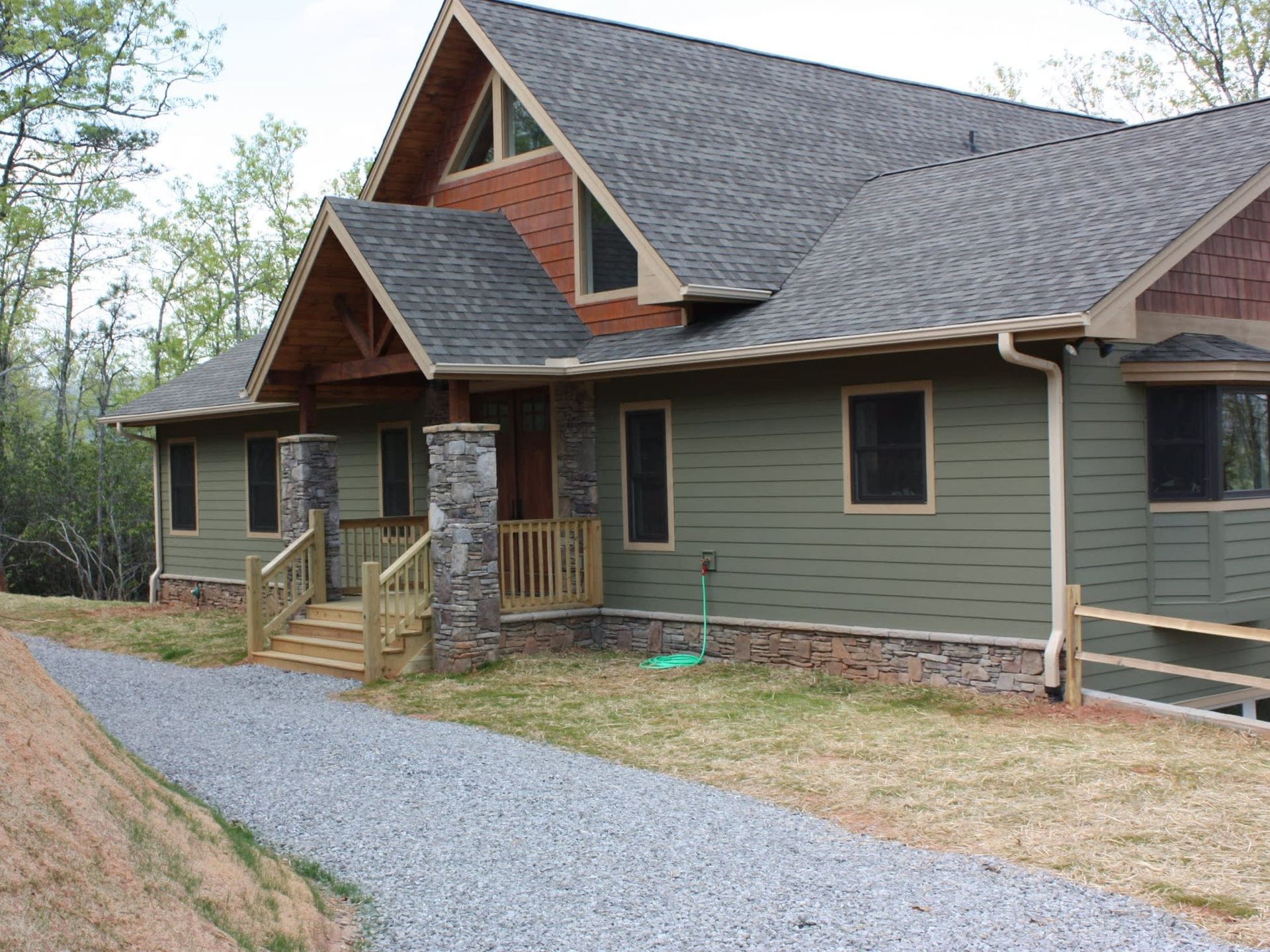 Log cabin-style house with green siding, stone accents, and a gravel driveway in a wooded setting
