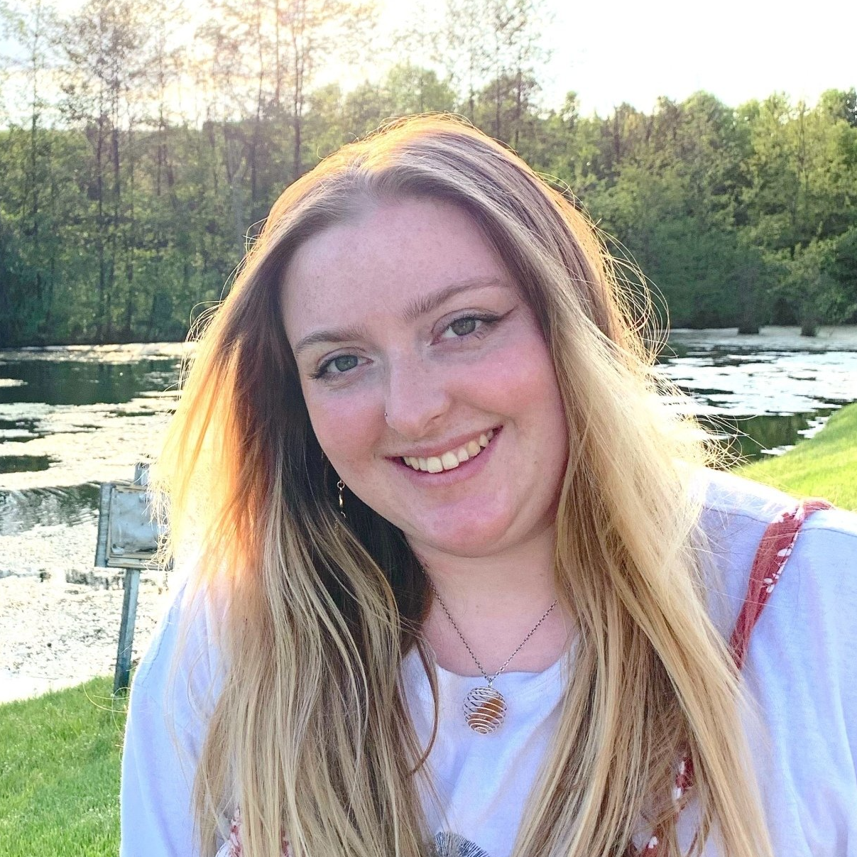 A woman with long blonde hair is smiling in front of a lake.