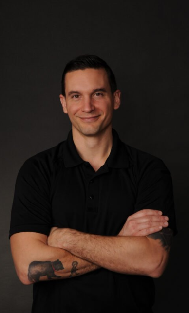 A man with a tattoo on his arm is standing with his arms crossed and smiling.