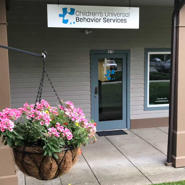 A sign for children 's universal behavior services hangs above a hanging basket of pink flowers