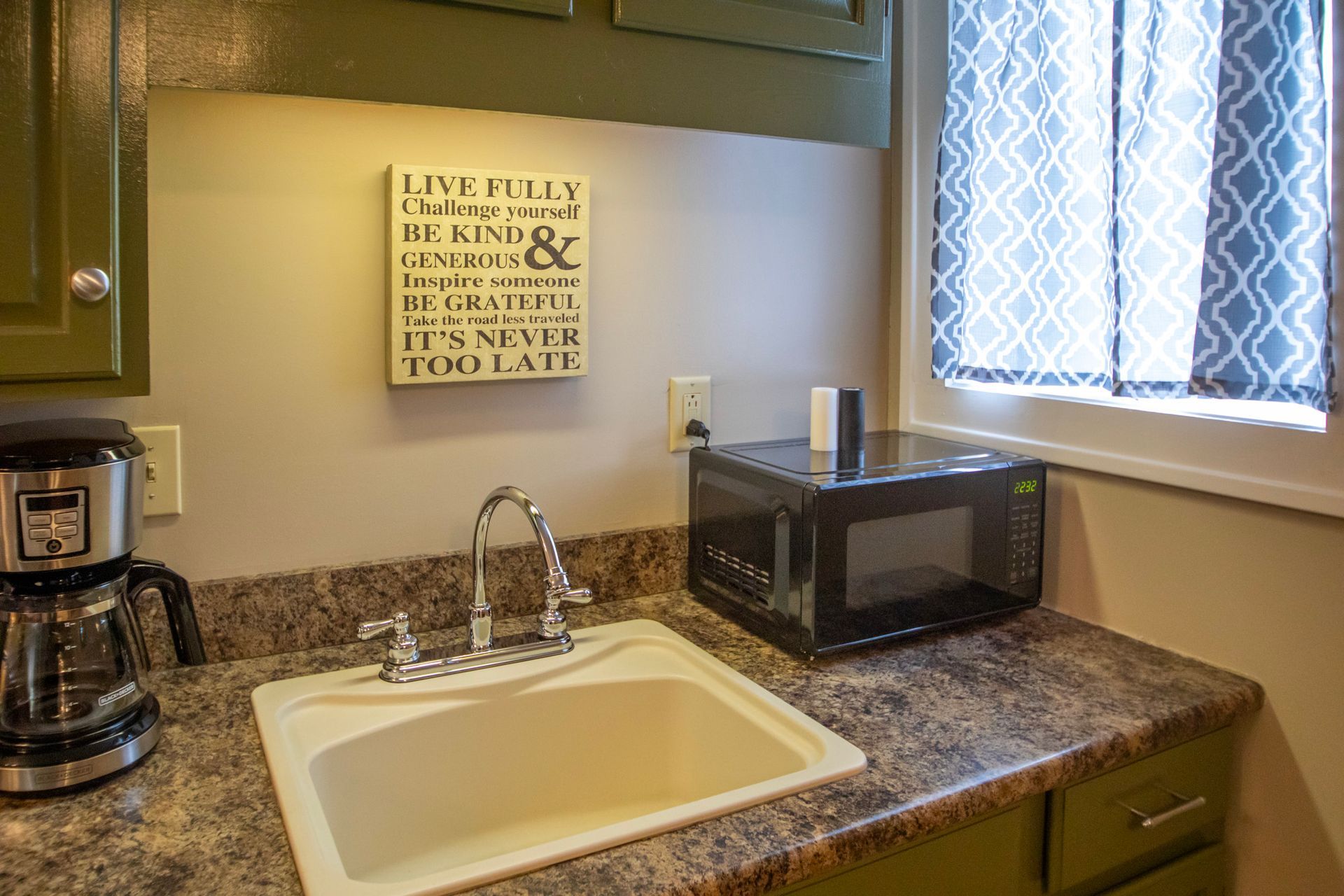 Kitchen counter with sink, microwave, and coffee maker. Green cabinets, window with patterned curtain, and sign on the wall.