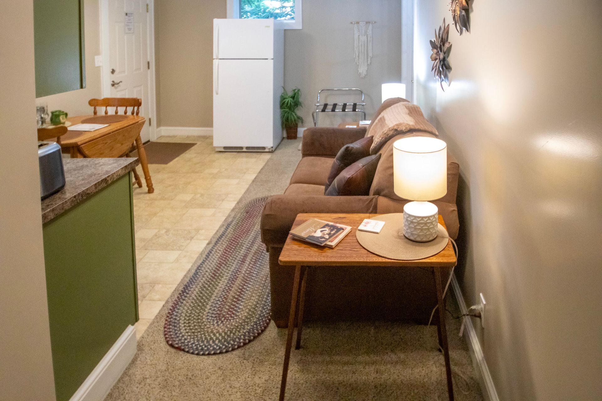 Cozy, narrow room with a brown couch, side table, small kitchen table, and rug. White refrigerator and off-white walls.