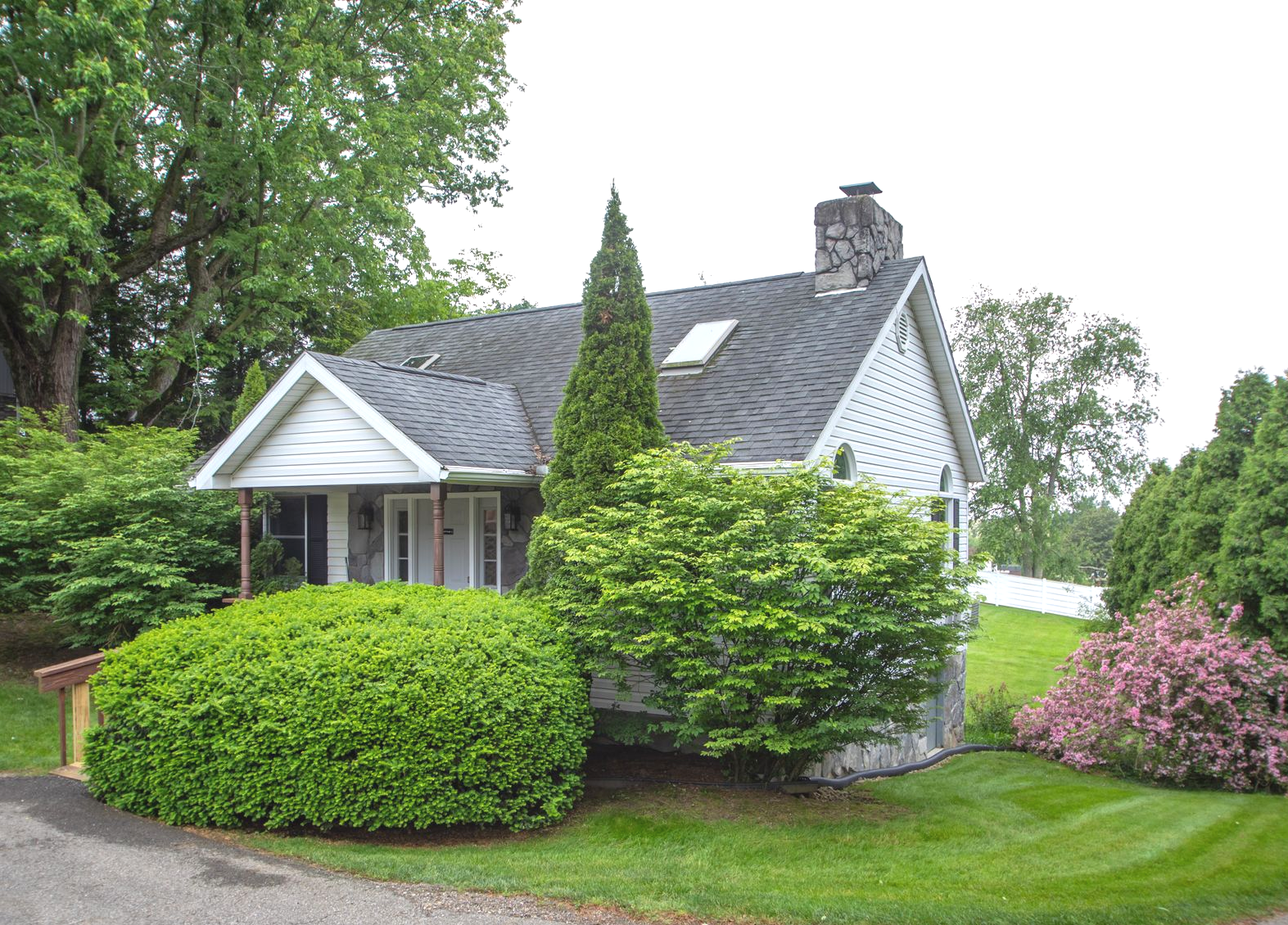 Small white house with dark gray roof, surrounded by green bushes and trees.