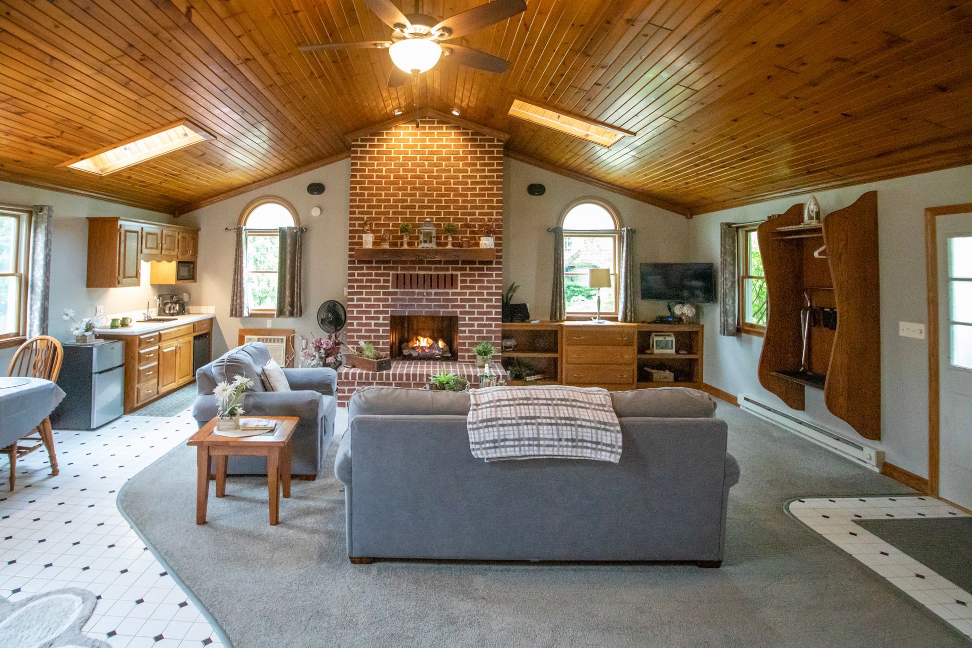 Cozy living room with fireplace, arched windows, and wood-paneled ceiling. Gray sofa, rug, and tiled floors.