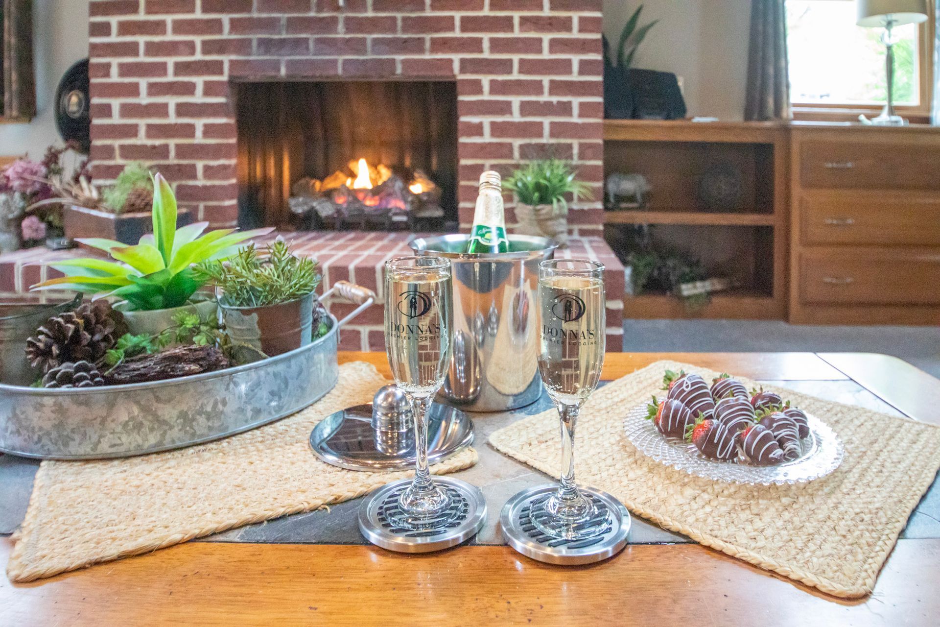 Champagne glasses, a bucket, and chocolate-covered strawberries on a table set for a celebration, with a lit fireplace.