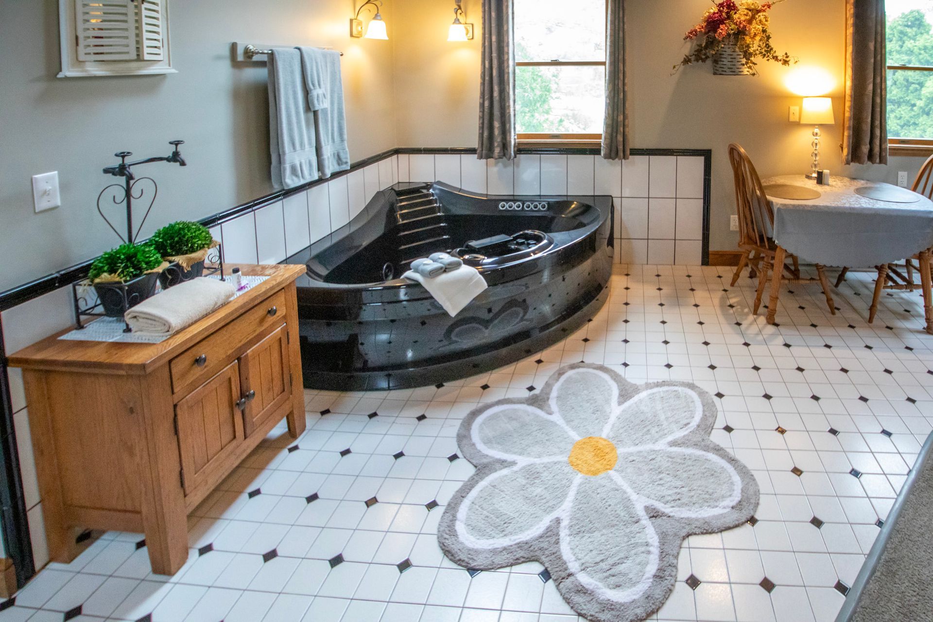 Bathroom with a black jacuzzi tub, wooden cabinet, daisy rug, and small dining table.