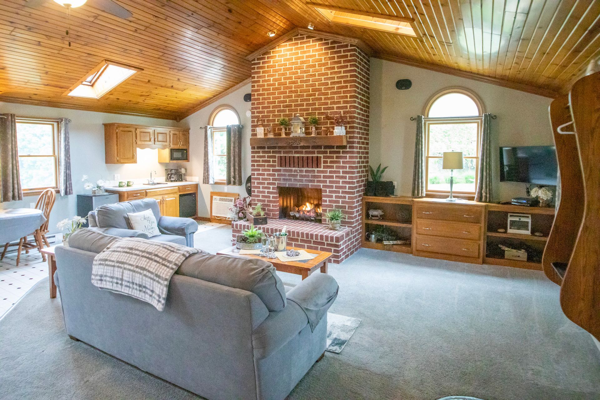Cozy living room with a brick fireplace, grey sofa, wooden ceiling and cabinets, and two arched windows.