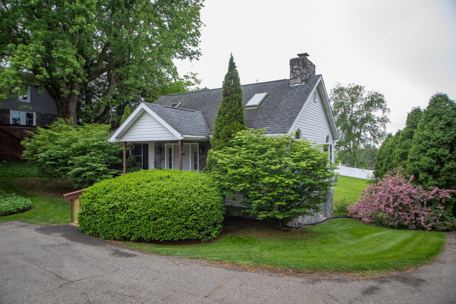 Small white house with dark roof surrounded by green bushes and trees, on a slight hill.