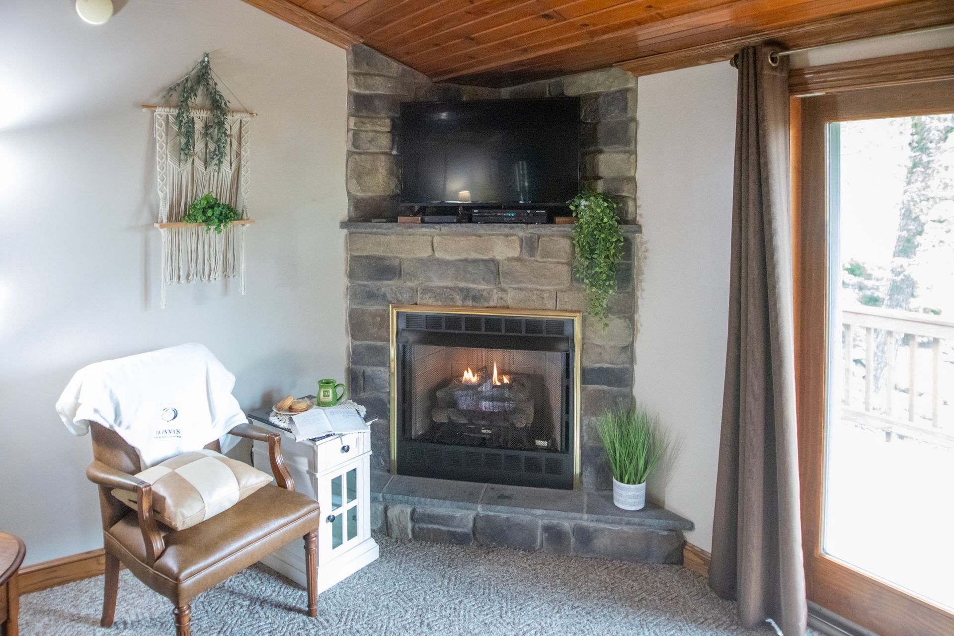 Cozy living room with fireplace, TV, armchair, macrame wall hanging, and wooden ceiling.