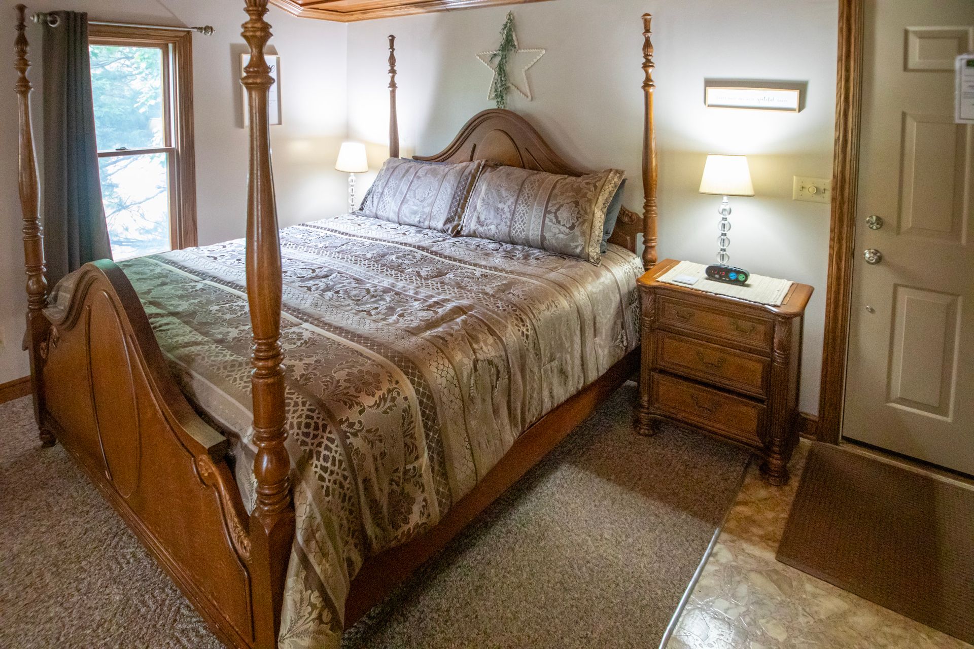 Bedroom with four-poster bed, nightstand, lamp, and window. Warm tones, beige carpet.