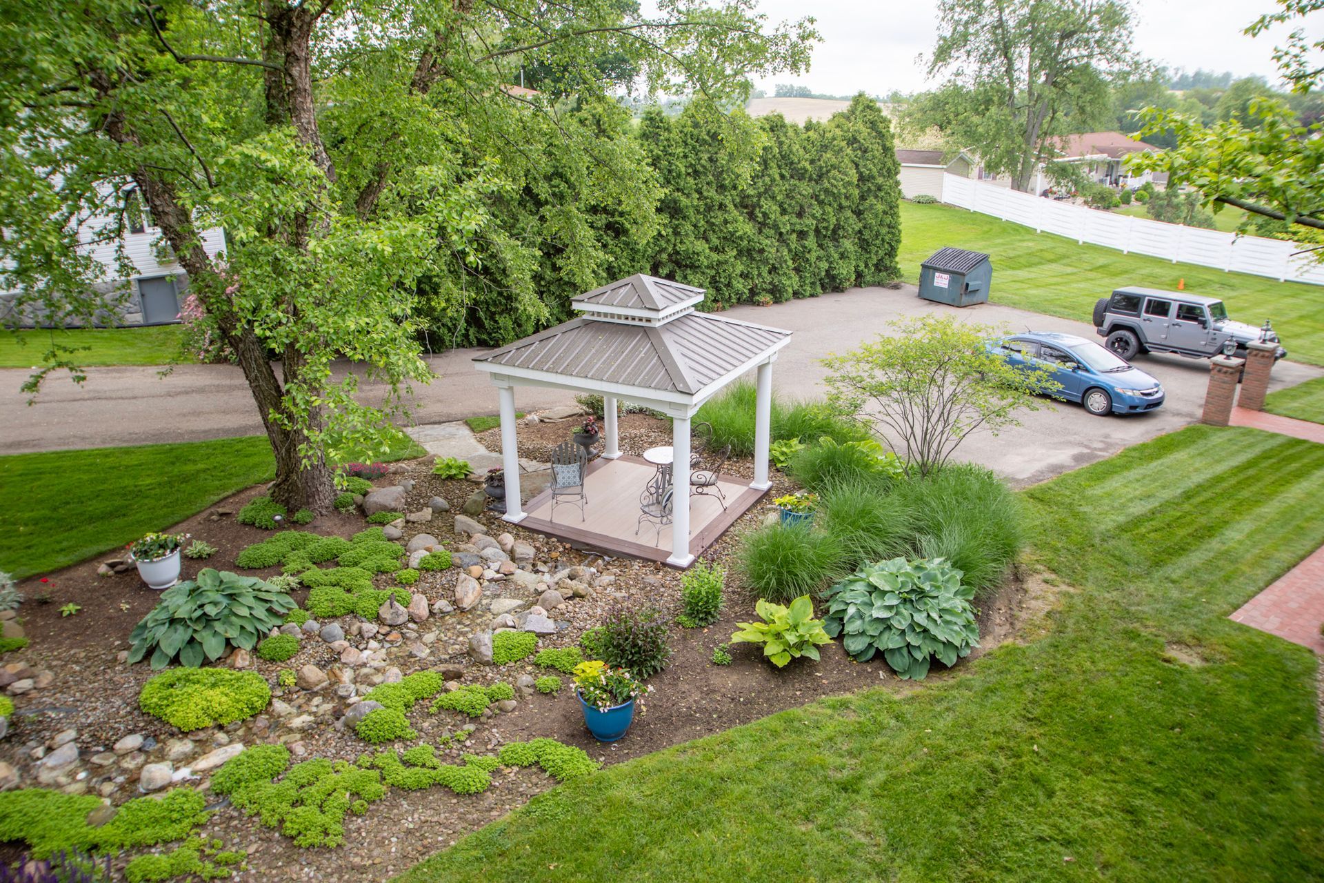 Gazebo in garden with plants and parked cars in background.