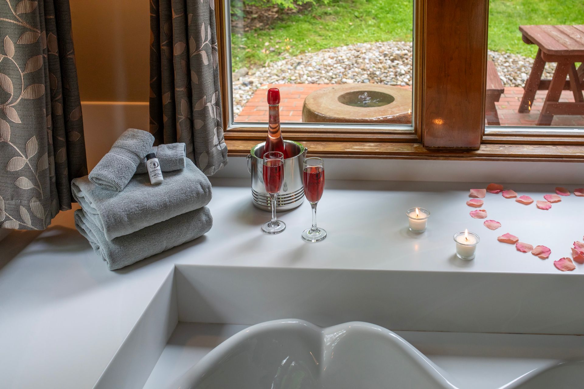 Bathtub scene: Champagne, candles, towels, rose petals in heart shape on a white ledge near a window overlooking a garden.