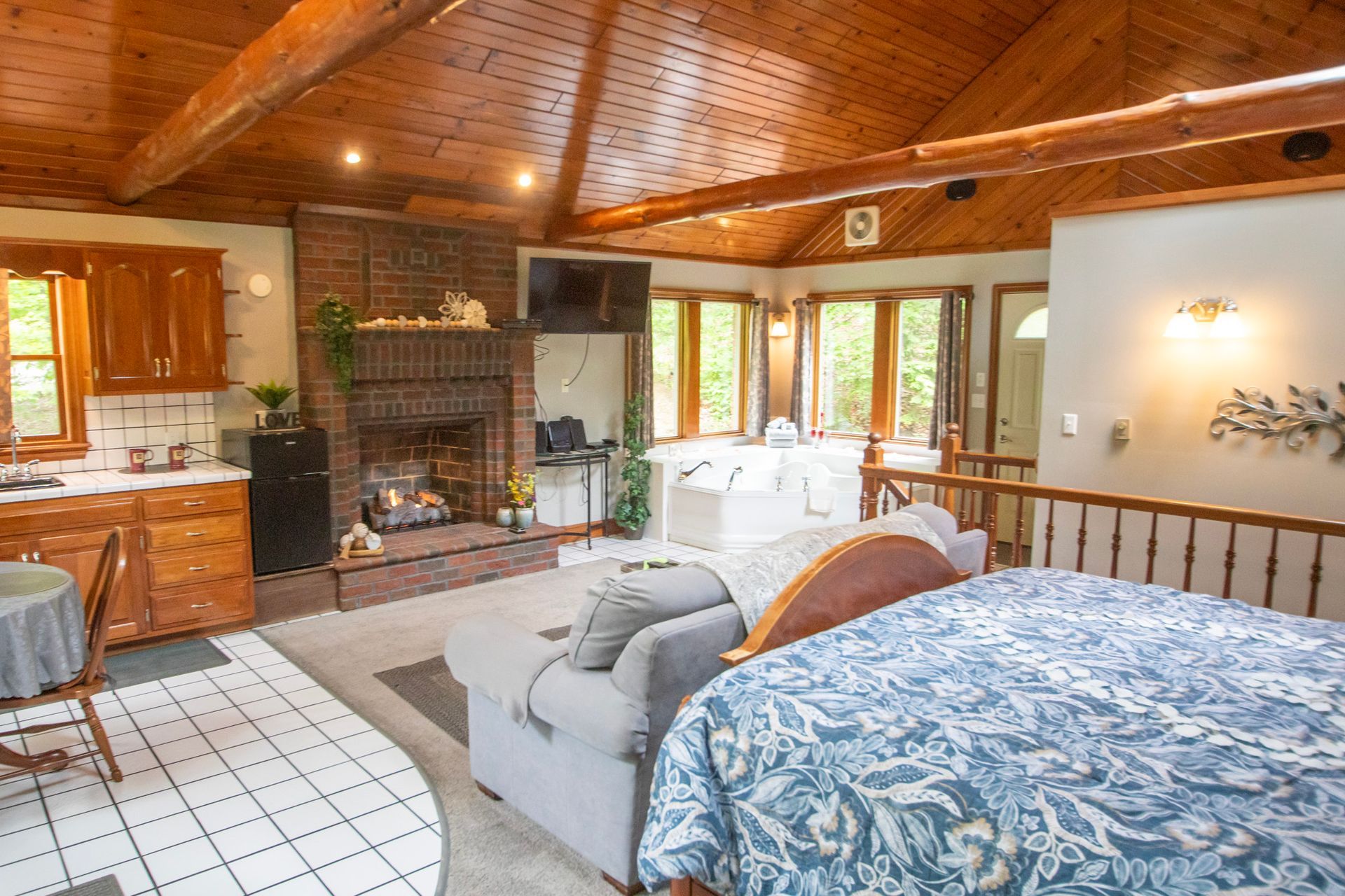 Open-plan living space with kitchen, fireplace, bed, and multiple windows; wood ceiling and beams; neutral color scheme.