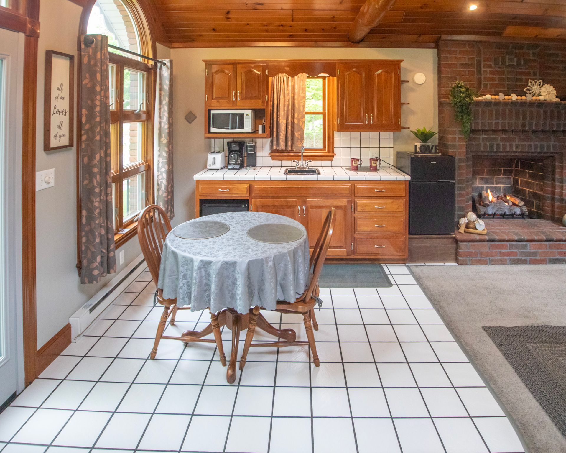 A small kitchen area with a table, chairs, cabinets, a fridge, and a fireplace. White tiled floor.