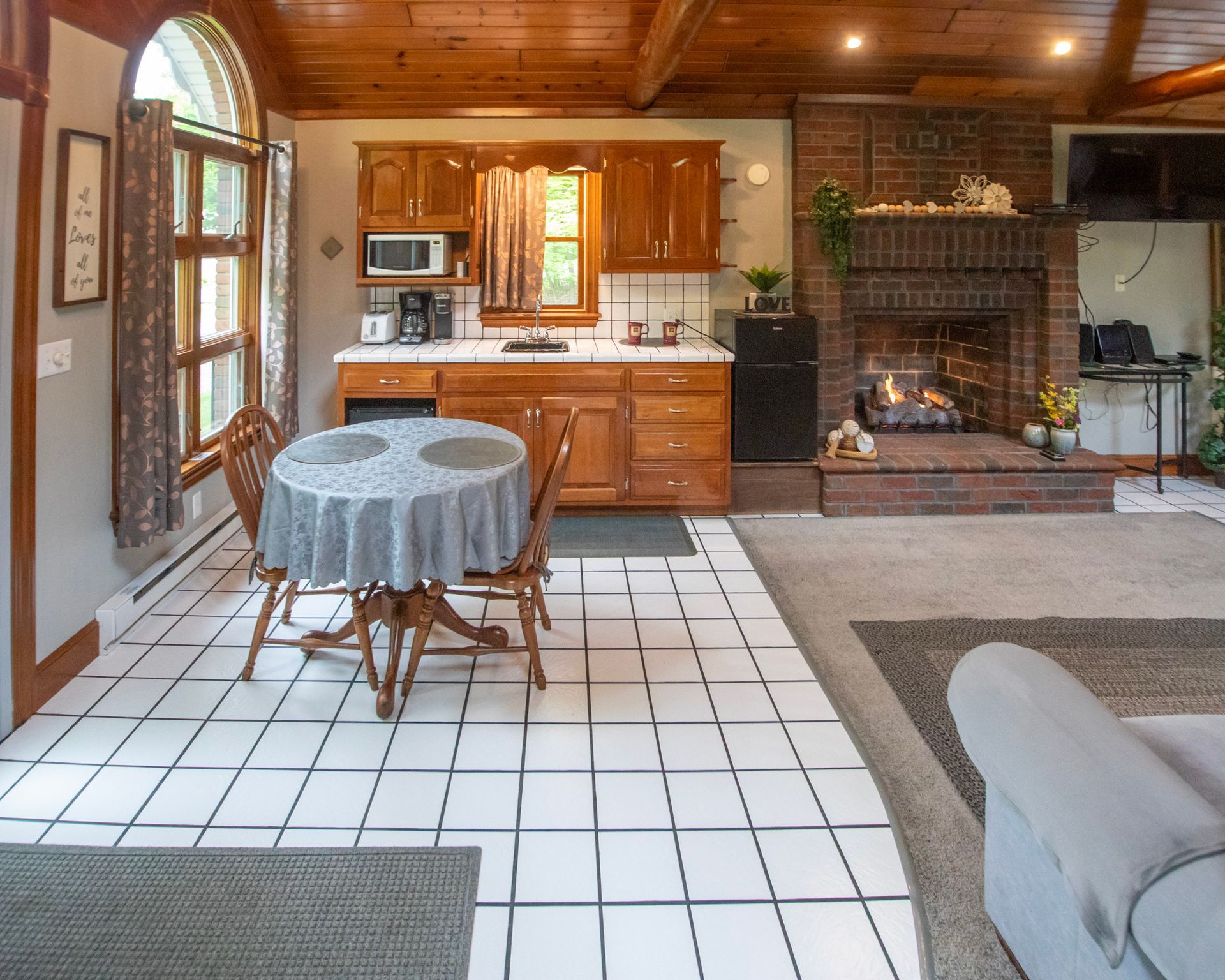 Kitchen and dining area with fireplace. Wooden cabinets, round table with a tablecloth, and white tiled floor.