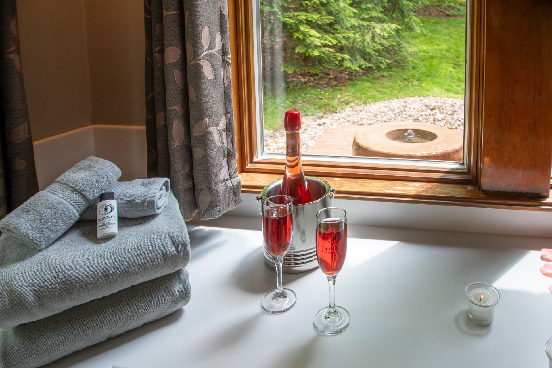 A bathtub with towels, champagne, and a view of a green backyard.